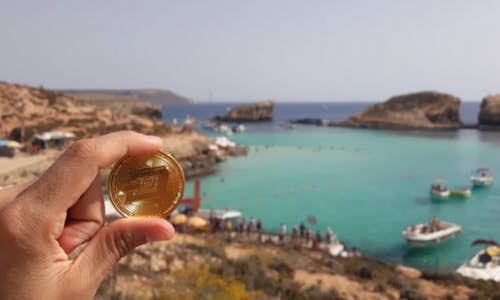 A photo of the coastline somewhere in Greece with turquoise seas. In the forefront, someone is holding a Cryptocurrency coin in front of the camera